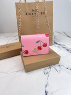 A pink Coach crossbody bag with a cherry print and gold chain strap, displayed on a brown Coach box with a Coach shopping bag in the background.