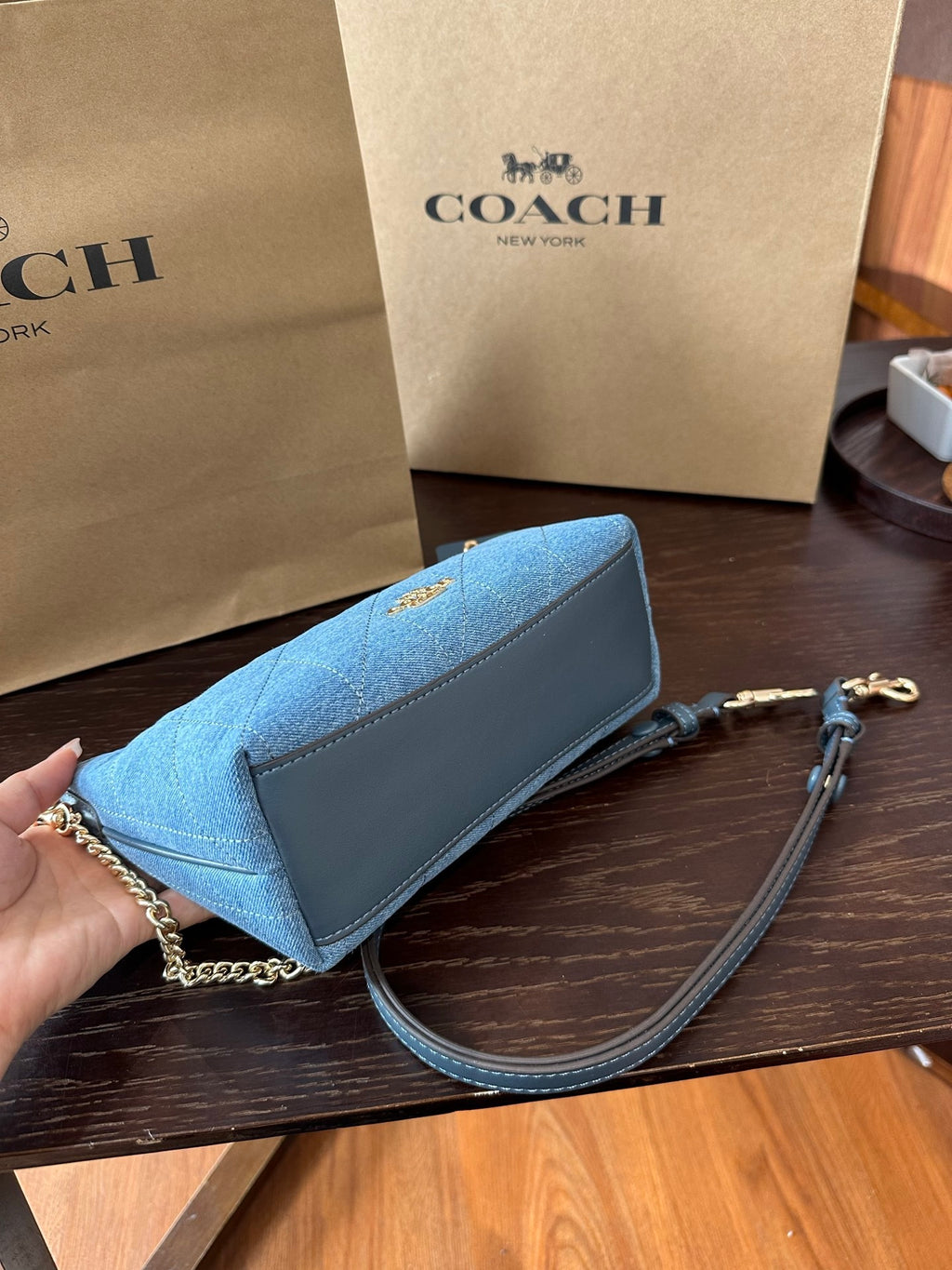 A Coach handbag in blue leather and canvas, held by a hand on a dark wooden table with Coach shopping bags in the background.