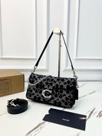 A Coach shoulder bag in black coated canvas with the signature monogram pattern covered in silver rhinestones, displayed on a stand with its strap, dust bag, and box on a marble surface.