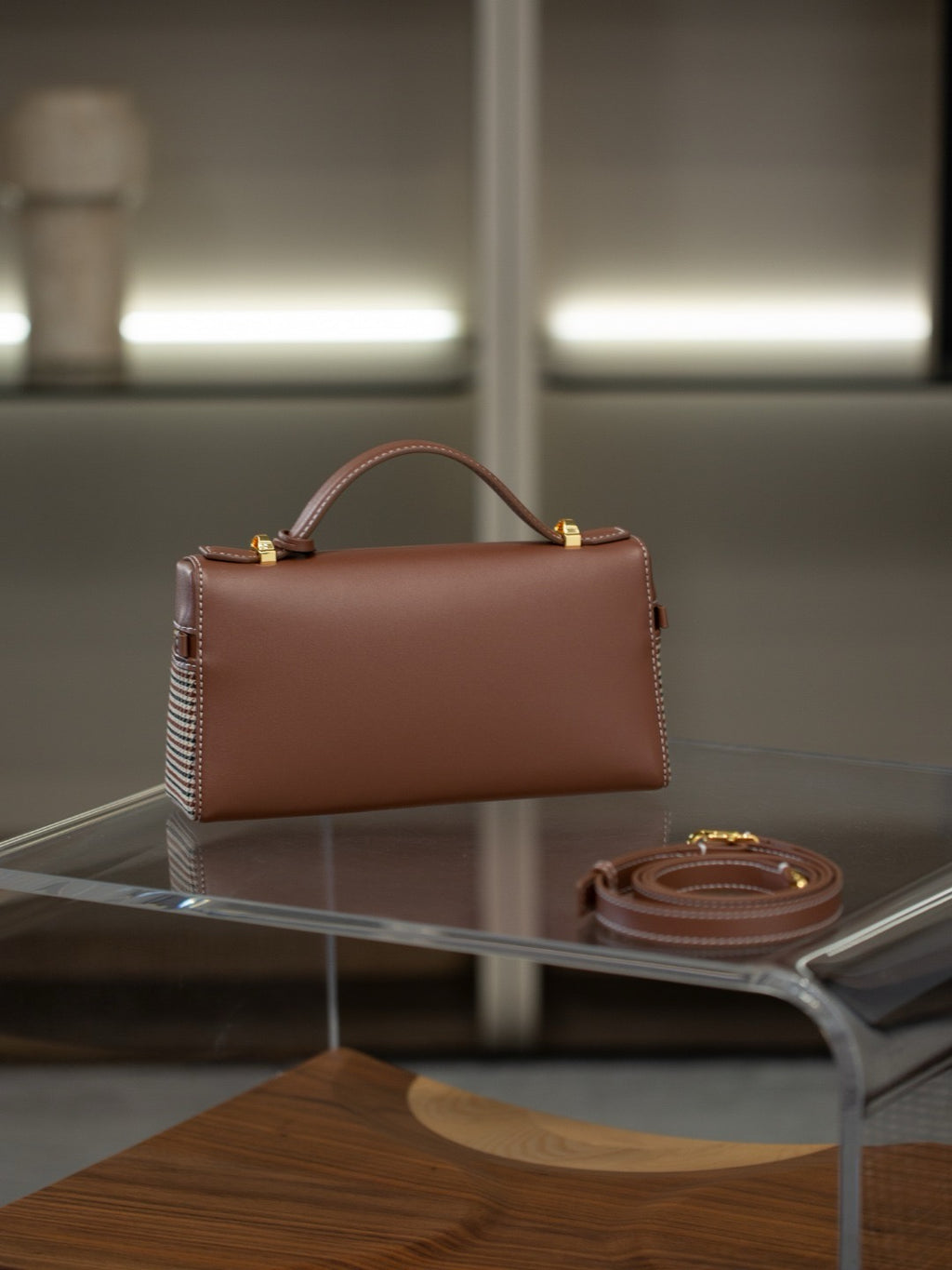 A structured brown leather handbag with a top handle and gold-tone hardware, displayed on a clear acrylic table with its matching strap beside it.