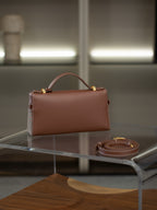 A structured brown leather handbag with a top handle and gold-tone hardware, displayed on a clear acrylic table with its matching strap beside it.