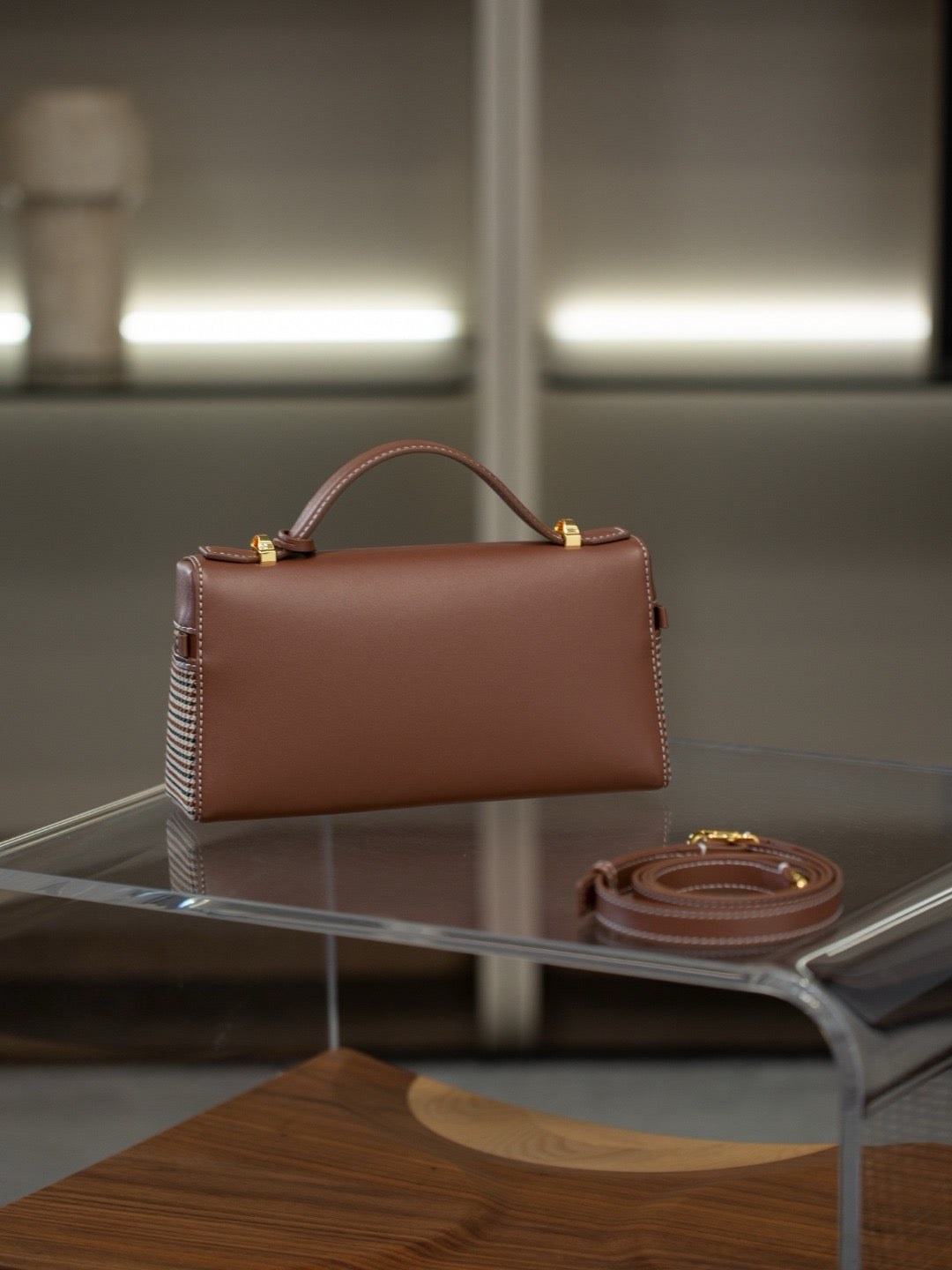 A structured brown leather handbag with a top handle and gold-tone hardware, displayed on a clear acrylic table with its matching strap beside it.