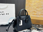 A black Coach handbag with top handles and a shoulder strap, displayed on a marble surface with a Coach shopping bag and box in the background.