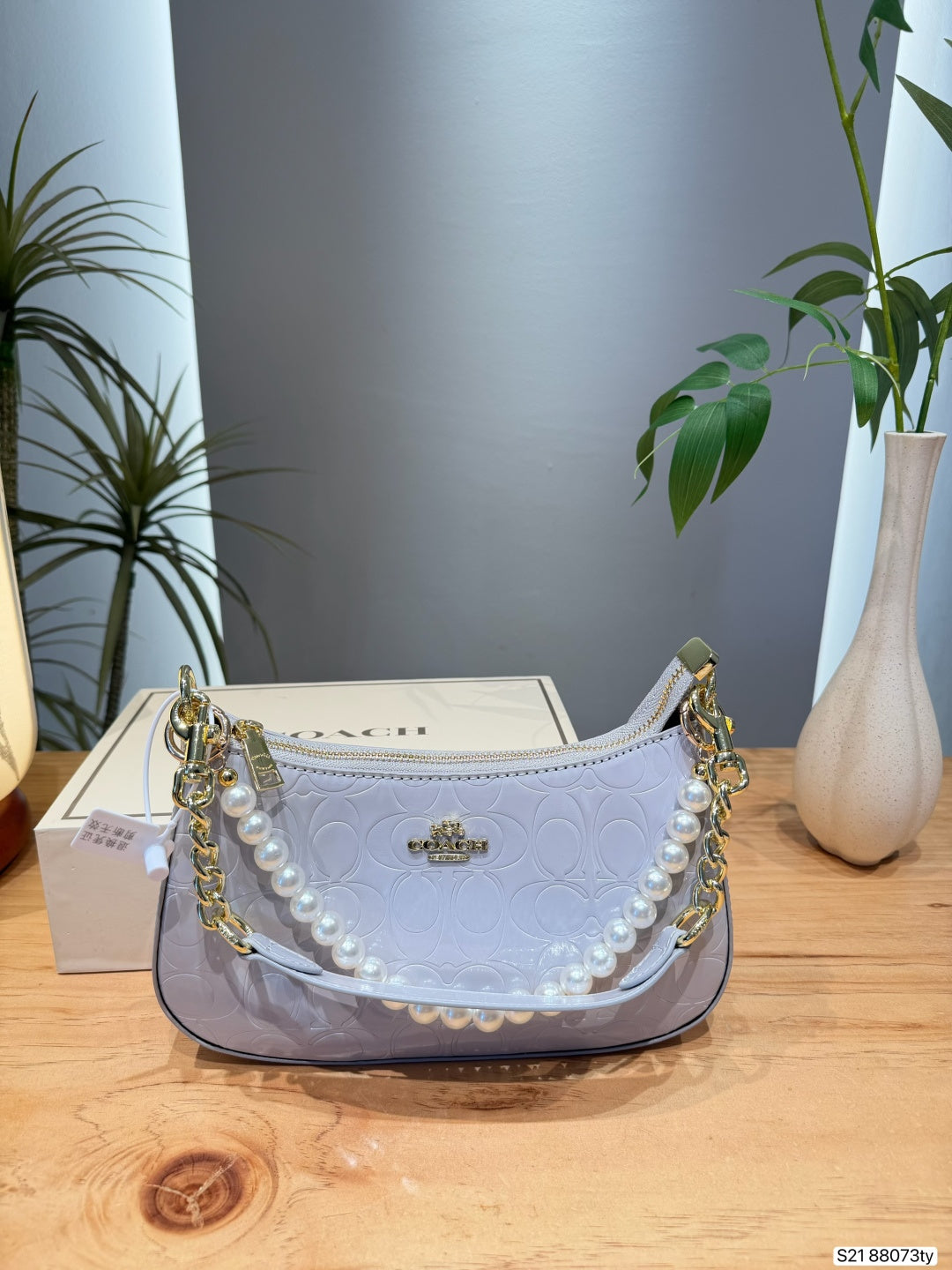 A Coach crescent-shaped shoulder bag in pale lavender, featuring an embossed logo pattern, a pearl-bead strap, and gold-tone chain details, displayed on a wooden table with a Coach box and plants in the background.