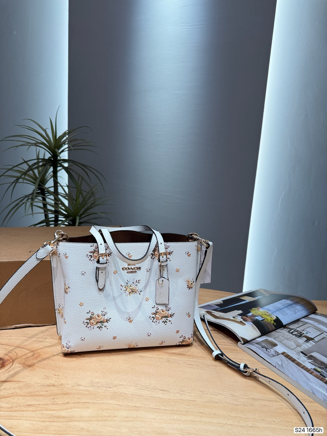 A white Coach handbag with a delicate floral pattern, displayed on a light wooden table next to an open magazine. The bag features silver-tone hardware and a matching white shoulder strap.