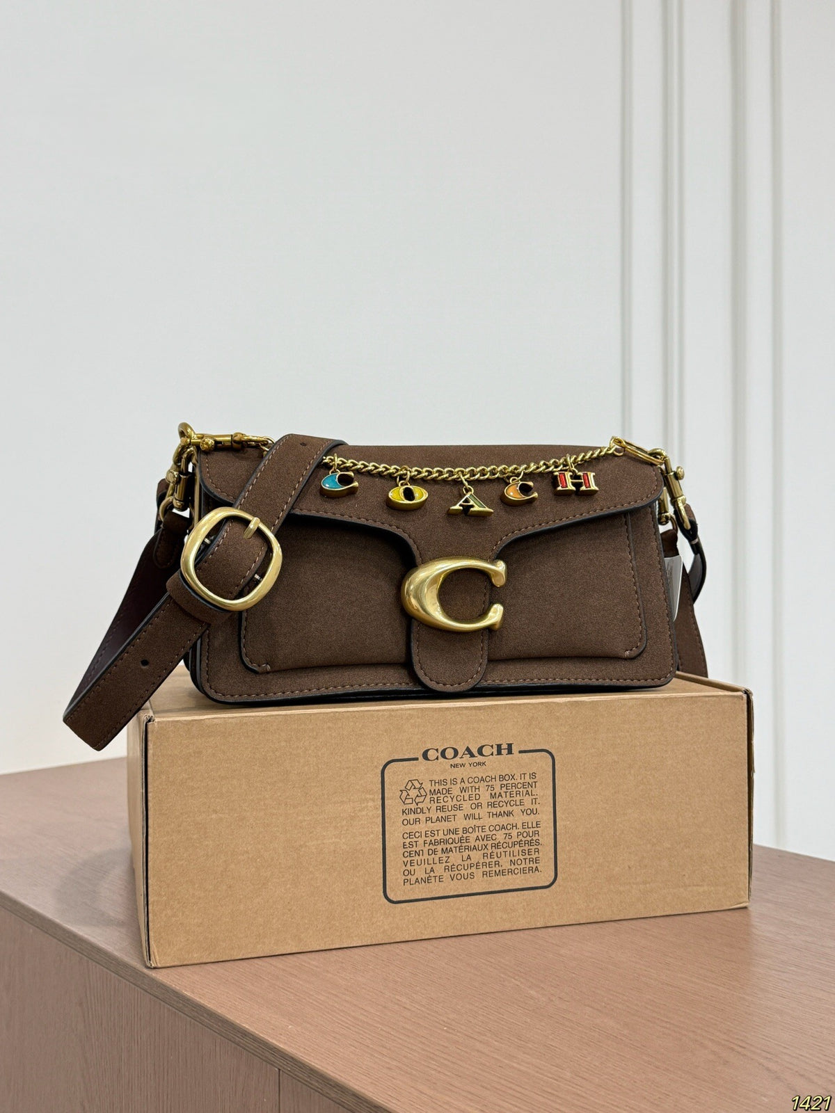 A Coach shoulder bag in dark brown suede and leather, featuring gold-tone hardware, a signature 'C' clasp, and colorful letter charms spelling 'COACH' on a chain strap. The bag is displayed on top of its original brown cardboard box on a wooden table against a white paneled wall.