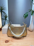 A gold Coach handbag with a signature C pattern, a pearl bead strap, and gold chain details, displayed on a wooden table with a Coach box and plants in the background.