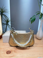 A gold Coach handbag with a signature C pattern, a pearl bead strap, and gold chain details, displayed on a wooden table with a Coach box and plants in the background.