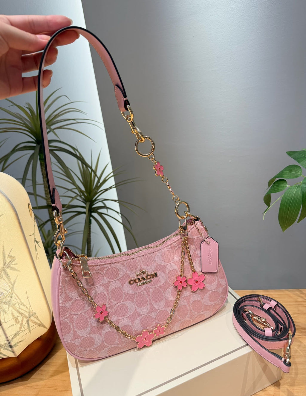 A pink Coach shoulder bag with a signature monogram pattern, gold-tone hardware, and pink flower charms, shown with a hand holding the strap and an additional strap coiled beside it on a tabletop.