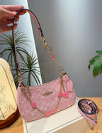 A pink Coach shoulder bag with a signature monogram pattern, gold-tone hardware, and pink flower charms, shown with a hand holding the strap and an additional strap coiled beside it on a tabletop.