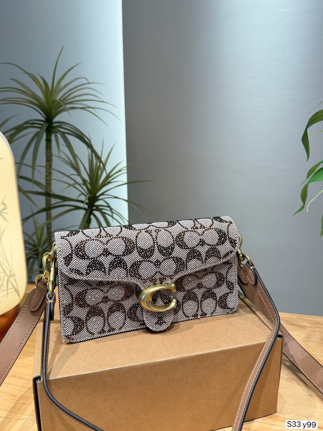 A Coach shoulder bag in the signature monogram canvas, resting on a brown cardboard box. The bag features a beige and brown pattern with gold-tone hardware, including the signature 'C' turn-lock clasp.