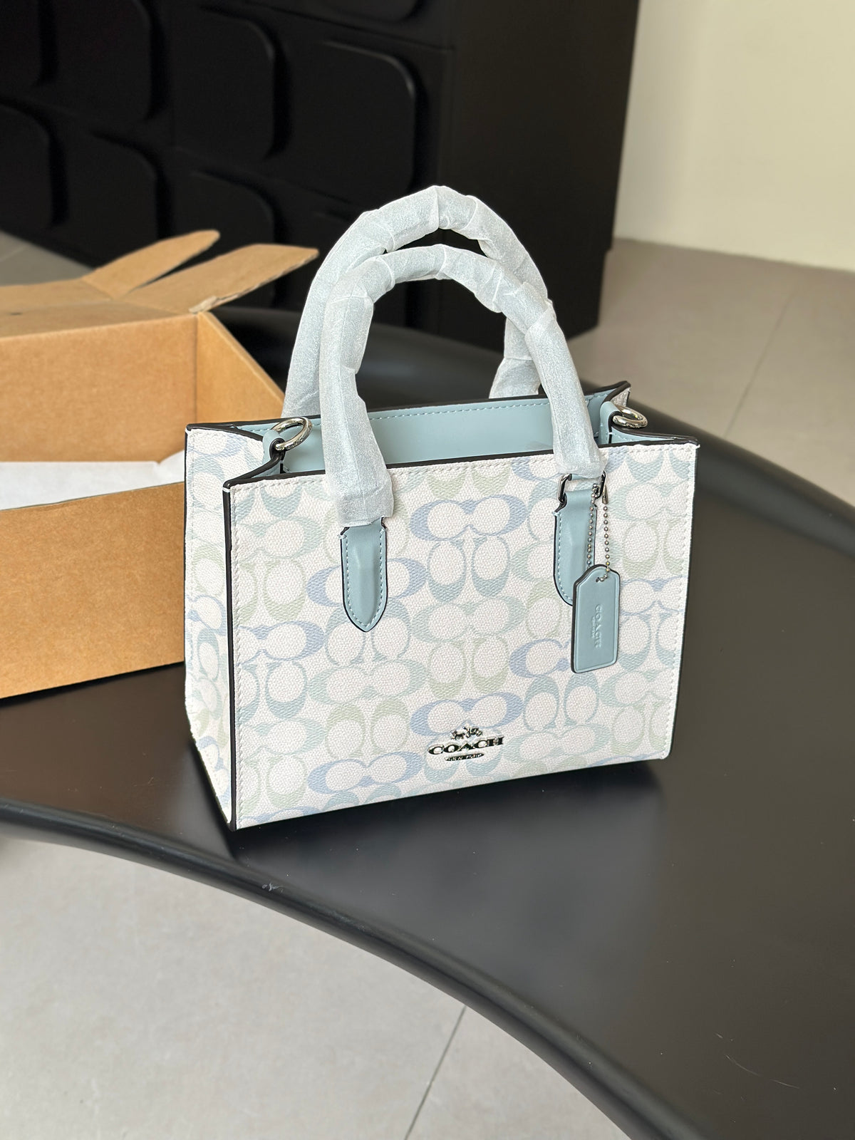 A Coach mini tote bag in the signature monogram canvas, featuring light blue leather trim and handles, sitting on a black table with a cardboard box in the background.
