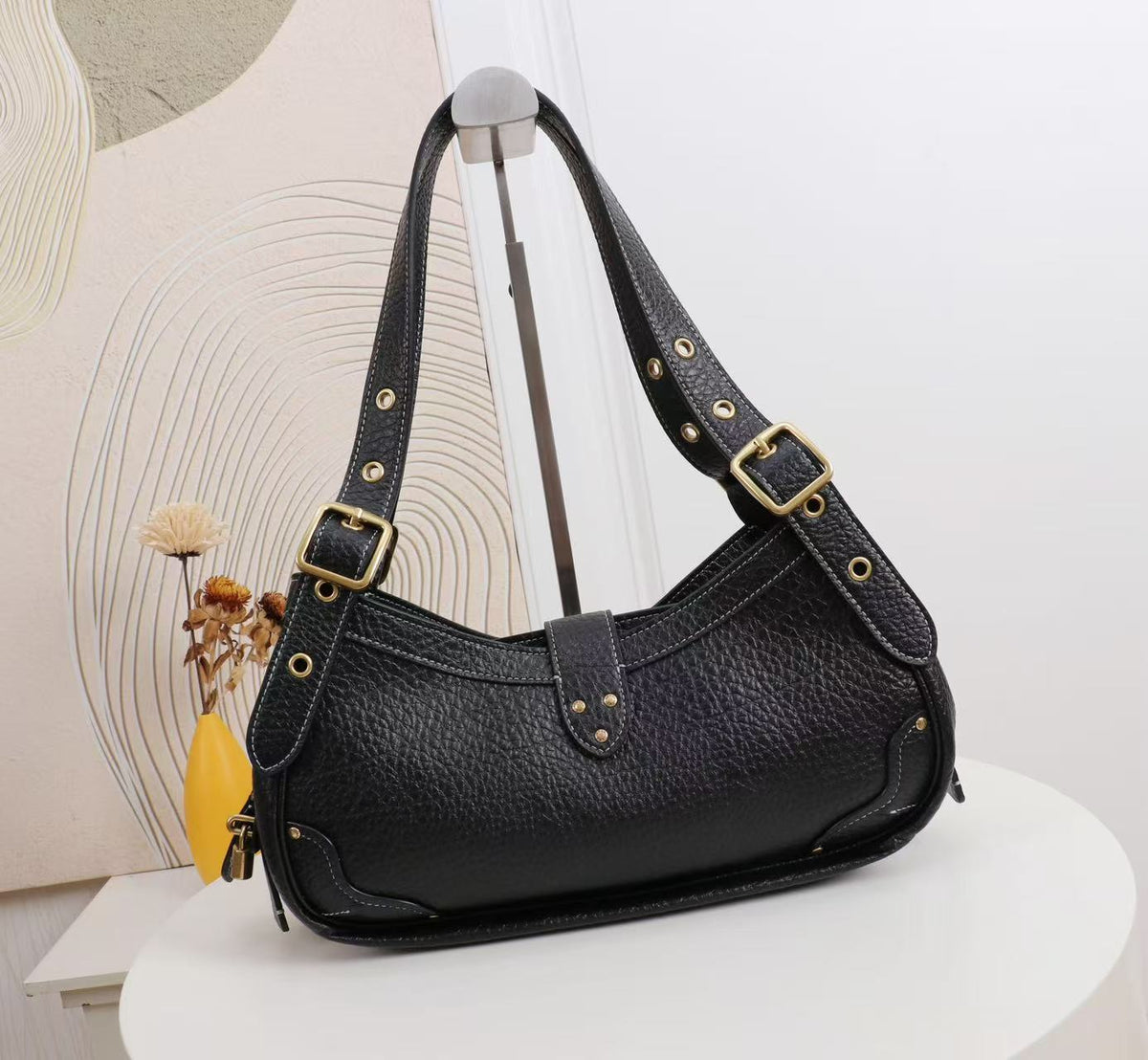 Black pebbled leather shoulder bag with gold-tone hardware, displayed on a white round table against a light beige wall with decorative dried flowers.
