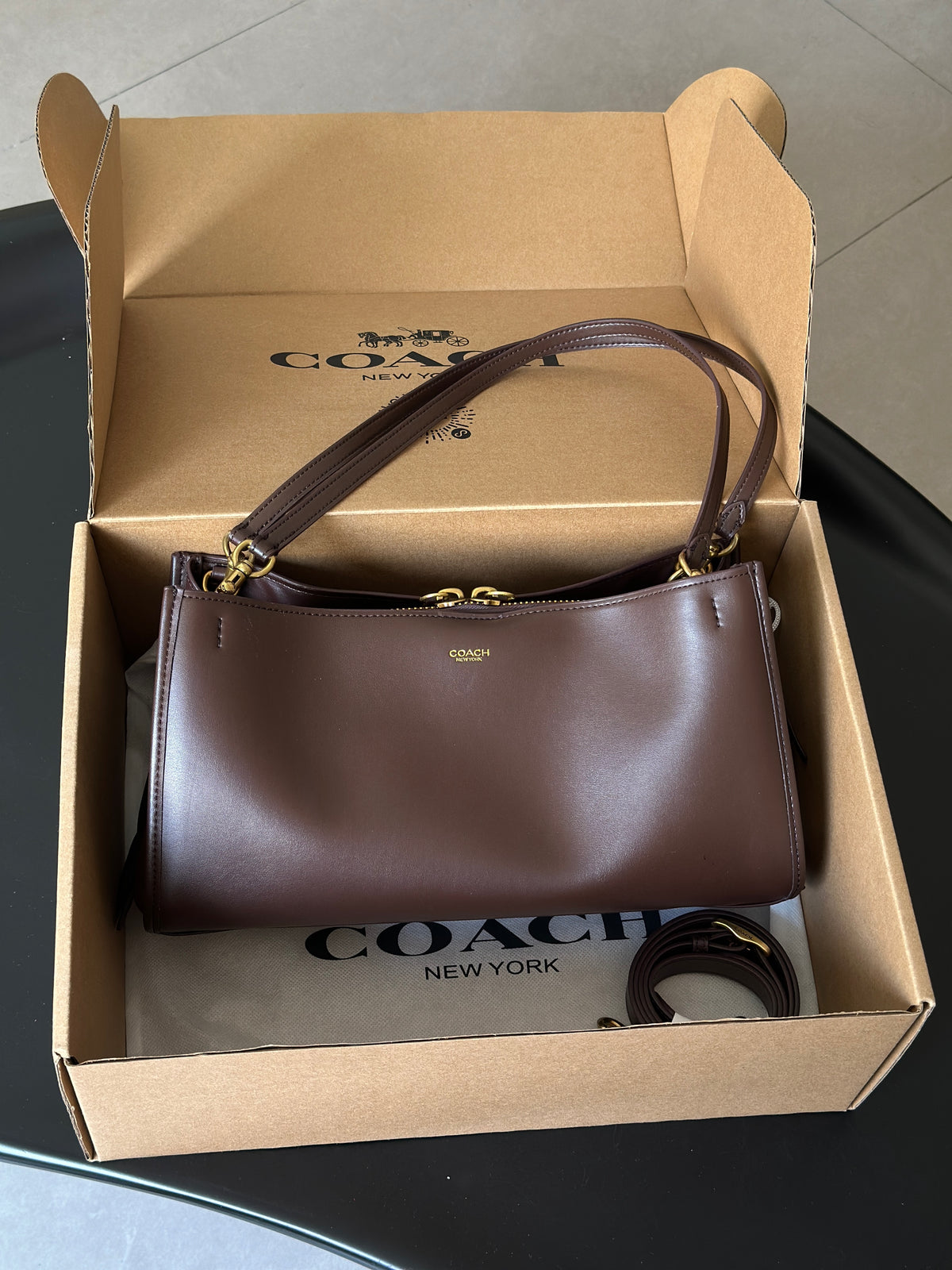 A Coach shoulder bag in dark brown leather with gold hardware, presented inside its open cardboard Coach box with the brand name visible on the box interior and dust bag.