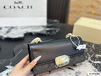 A Coach handbag with a black leather flap and signature monogram pattern base, held by a hand. The bag features gold-tone hardware and a small clear crystal accent on the front.