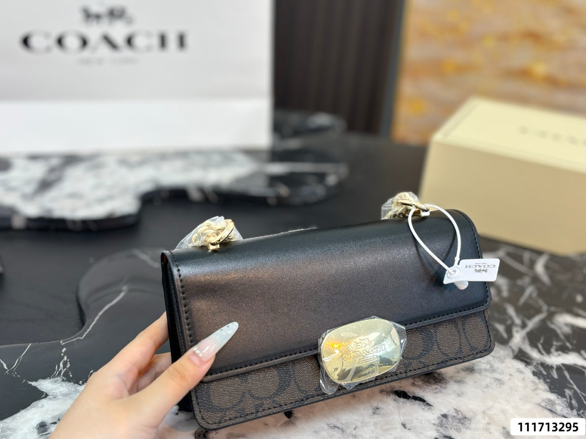 A Coach handbag with a black leather flap and signature monogram pattern base, held by a hand. The bag features gold-tone hardware and a small clear crystal accent on the front.