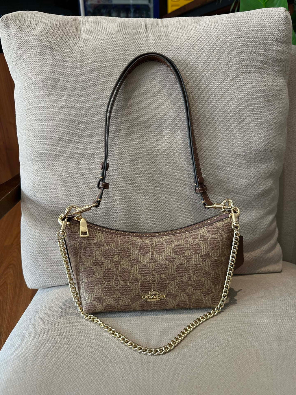 A Coach shoulder bag in brown signature monogram canvas with a gold-tone chain strap, resting on a beige upholstered chair in front of a large beige pillow.