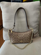 A Coach shoulder bag in brown signature monogram canvas with a gold-tone chain strap, resting on a beige upholstered chair in front of a large beige pillow.