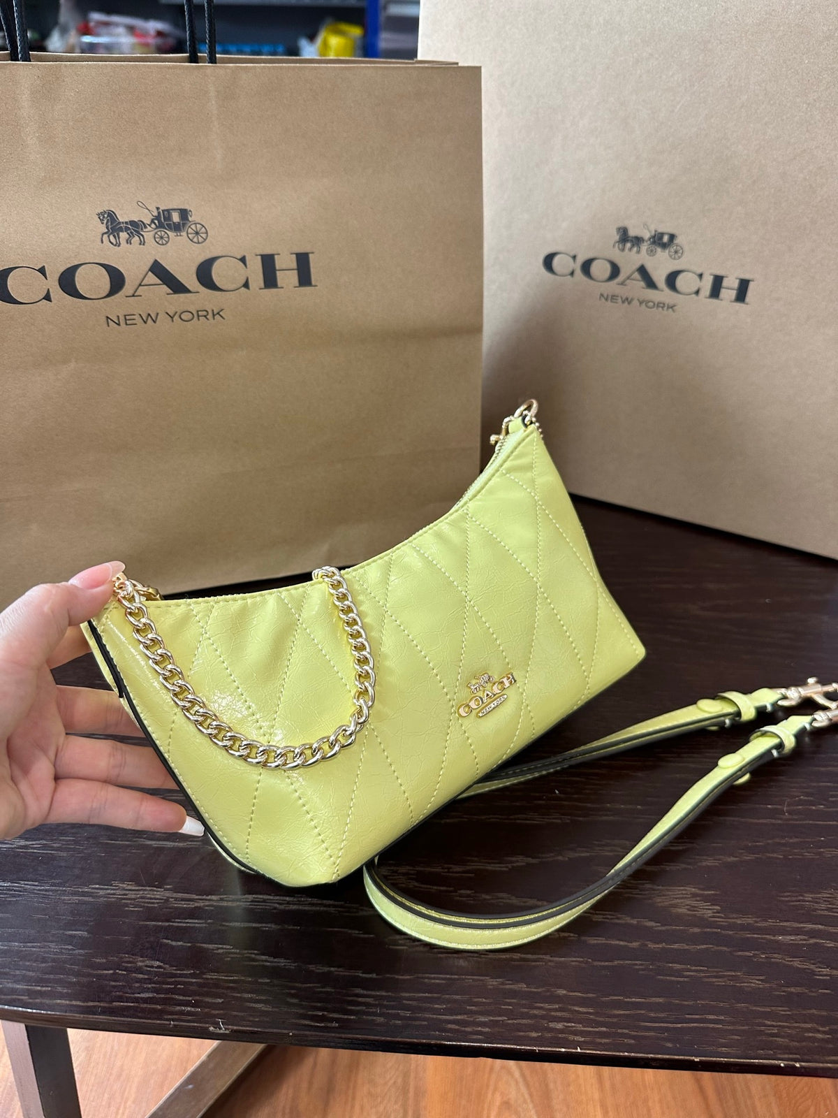 A Coach New York handbag in light yellow-green quilted leather, held by a hand in front of a brown Coach shopping bag on a dark wooden table.