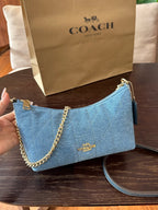 A Coach handbag in light blue quilted material with gold-tone chain and logo hardware, held by a hand in front of a Coach shopping bag on a dark wooden table.