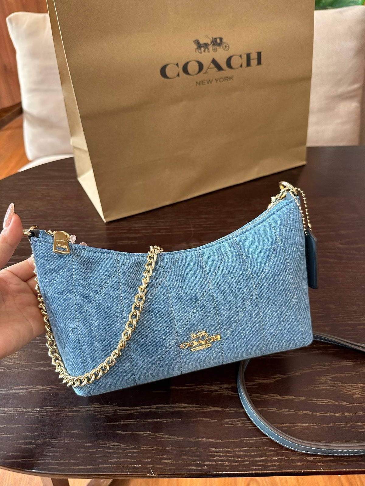 A Coach handbag in light blue quilted material with gold-tone chain and logo hardware, held by a hand in front of a Coach shopping bag on a dark wooden table.