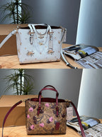 Two Coach handbags displayed side by side on a wooden table with a box and magazine in the background. The top bag is white textured leather with a floral print and gold-tone hardware.