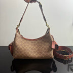 A Coach hobo shoulder bag in the signature monogram canvas, held by a hand. The bag features a tan and brown monogram pattern, reddish-brown leather trim, and gold-tone hardware.