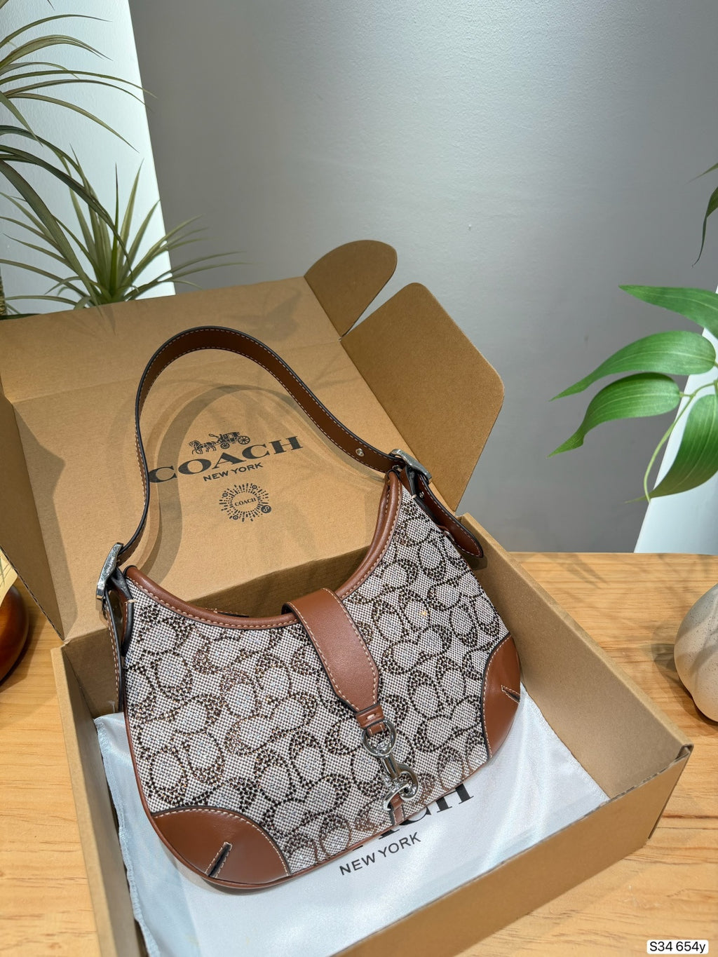 A Coach shoulder bag in the brand's signature monogram canvas and brown leather, displayed inside its open cardboard packaging on a white tissue paper, set on a wooden table with indoor plants in the background.
