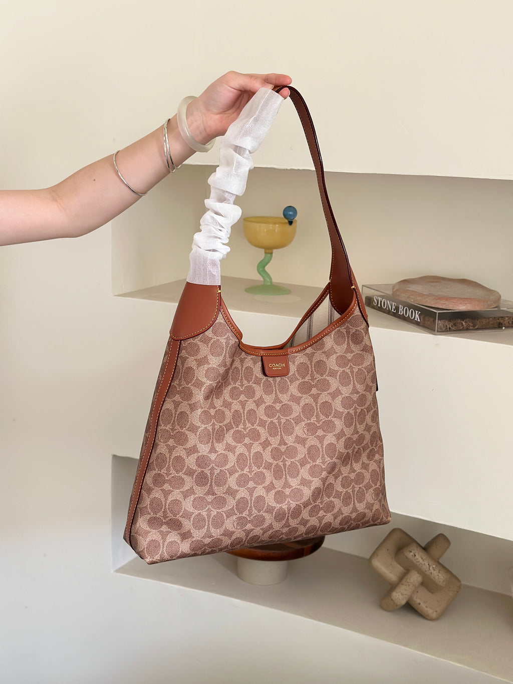 A person's hand holds up a Coach hobo shoulder bag with a brown and beige monogram pattern, featuring a distinctive white ruffled fabric strap, set against a minimalist beige shelving background.