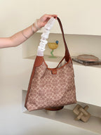 A person's hand holds up a Coach hobo shoulder bag with a brown and beige monogram pattern, featuring a distinctive white ruffled fabric strap, set against a minimalist beige shelving background.