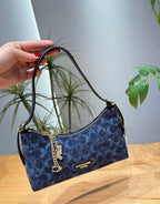A hand holds a Coach shoulder bag in navy blue signature C pattern canvas with gold hardware, set against a background of indoor plants and a wooden table.