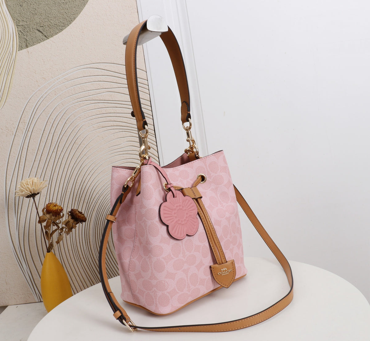 A pink Coach bucket bag with a signature monogram pattern, tan leather trim, a floral leather charm, and a long shoulder strap, displayed on a white round table.