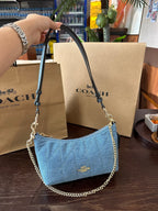 Blue handbag with chain detail held by a hand, with Coach bags and packaging in the background.