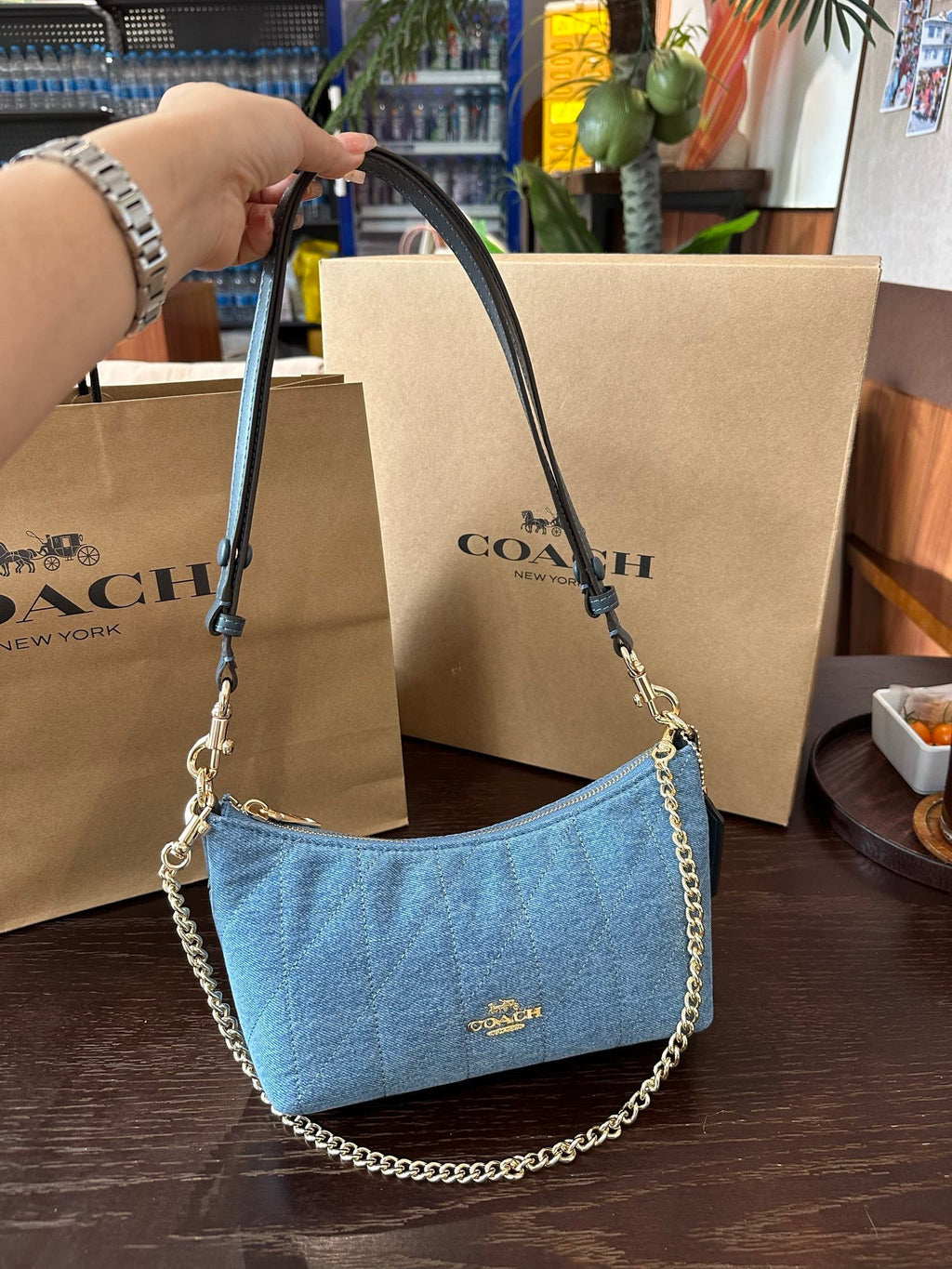 Blue handbag with chain detail held by a hand, with Coach bags and packaging in the background.