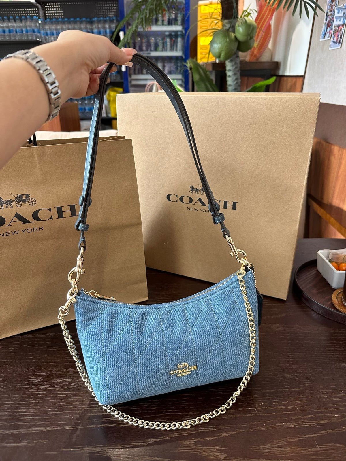 Blue handbag with chain detail held by a hand, with Coach bags and packaging in the background.