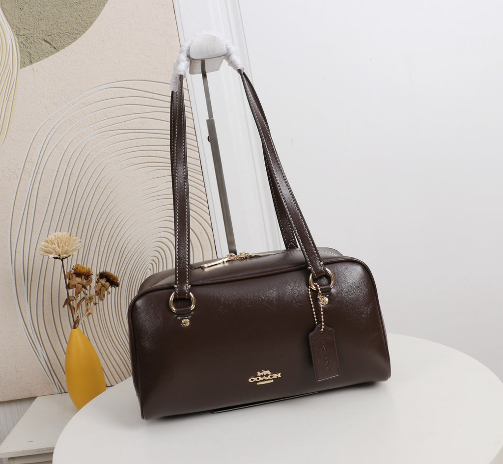 A dark brown Coach handbag with gold-tone hardware, displayed on a white round table against a white wall with a decorative abstract print and a yellow vase.