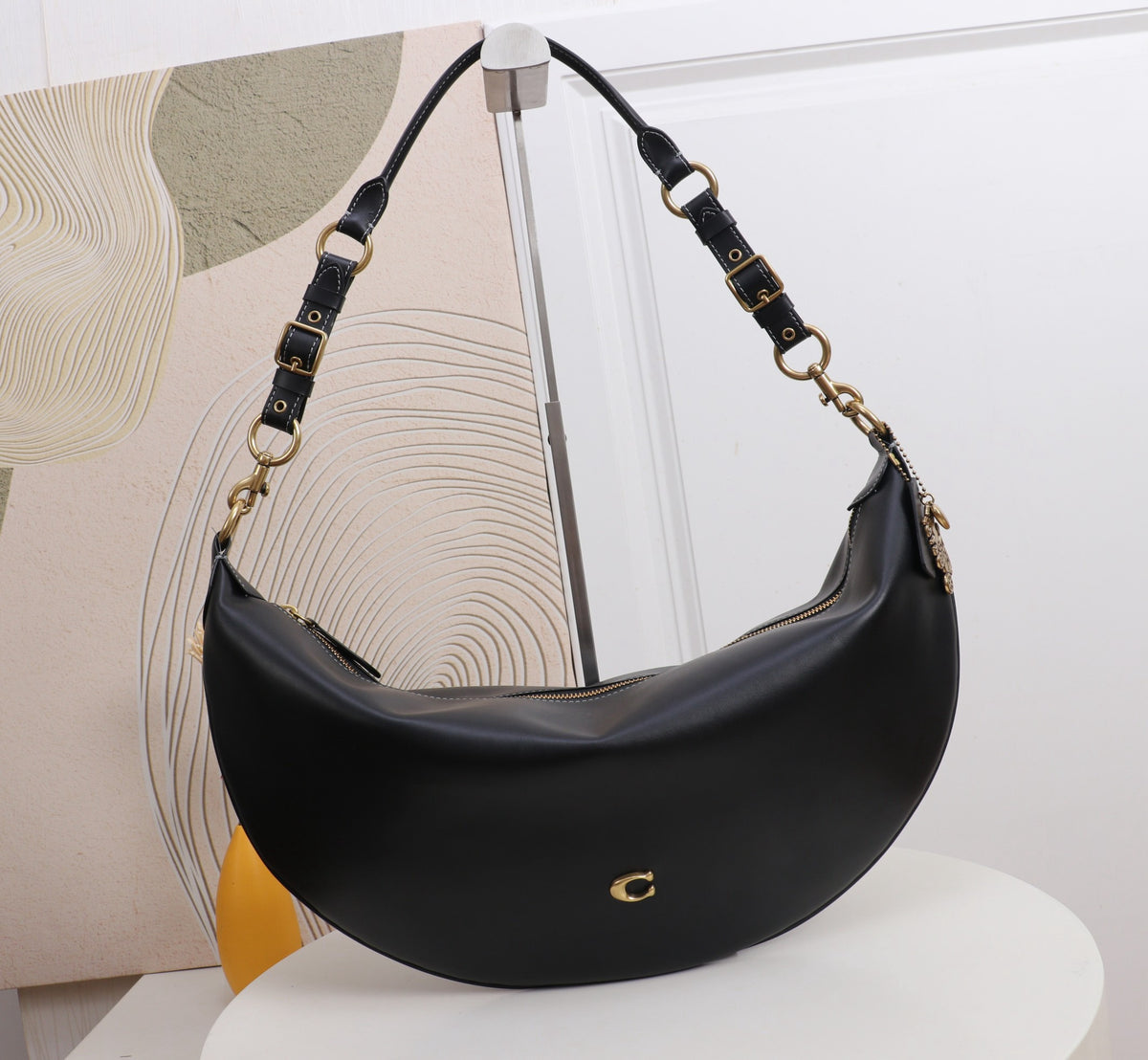 A black crescent-shaped leather shoulder bag with gold-tone hardware and a small gold logo on the front, displayed on a white pedestal in front of a modern abstract art piece.