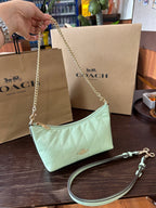 A Coach shoulder bag in pale mint green quilted leather, held by a hand. The bag features a gold-tone chain strap and a matching wristlet strap.
