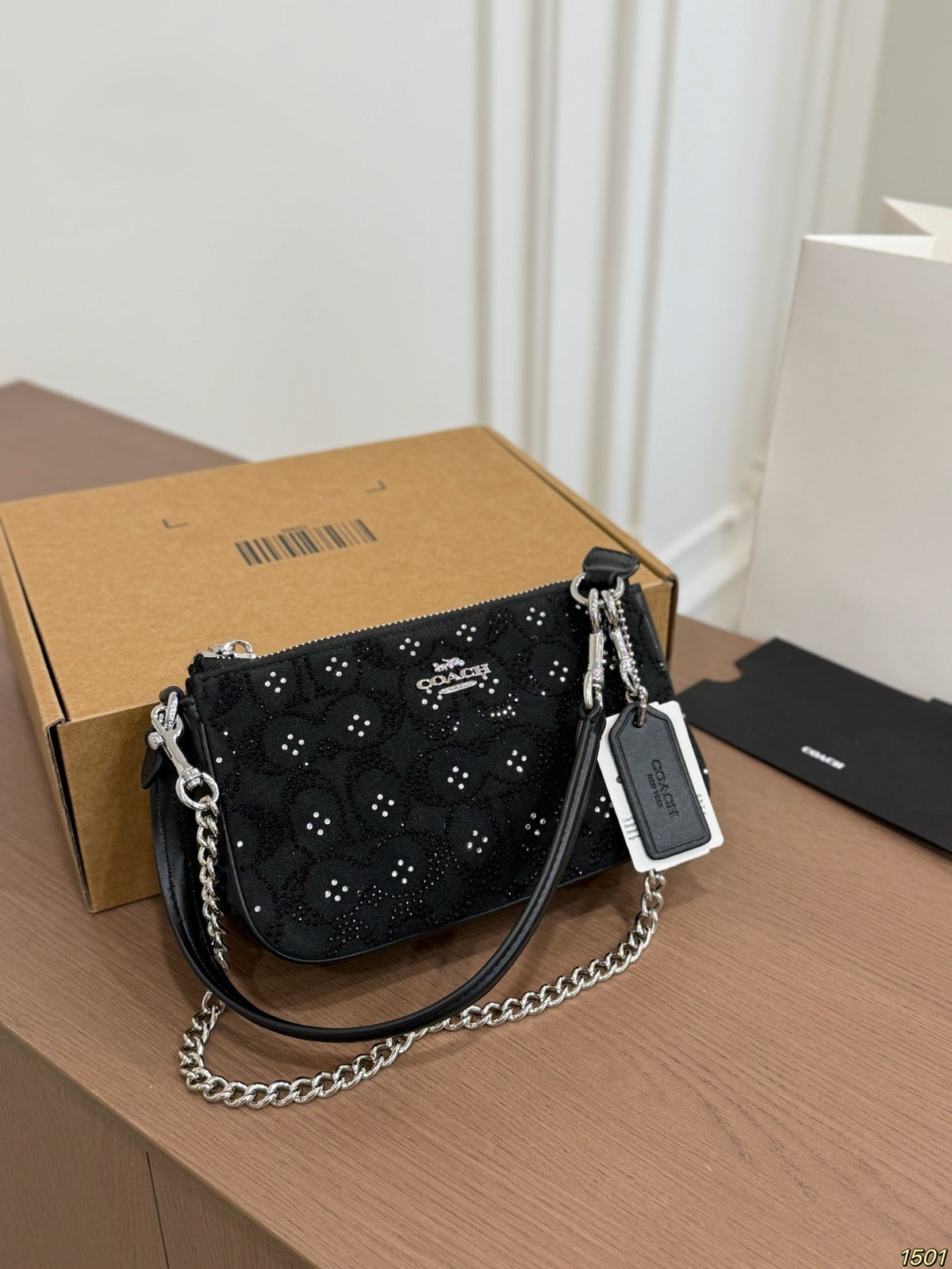 A black Coach shoulder bag with floral embroidery and silver-tone chain strap, resting on a wooden table next to its brown cardboard box.