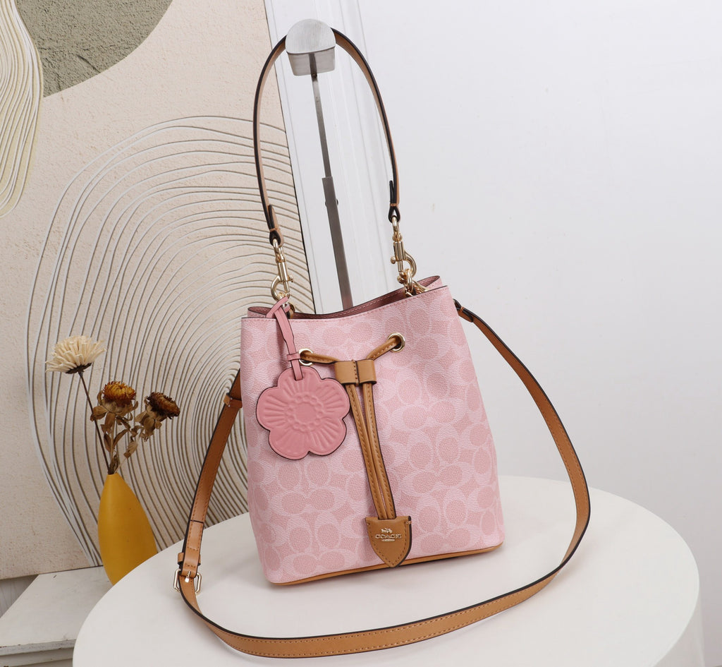 A pink Coach bucket bag with a monogram pattern, tan leather trim, a flower-shaped charm, and a long shoulder strap, displayed on a white round table.