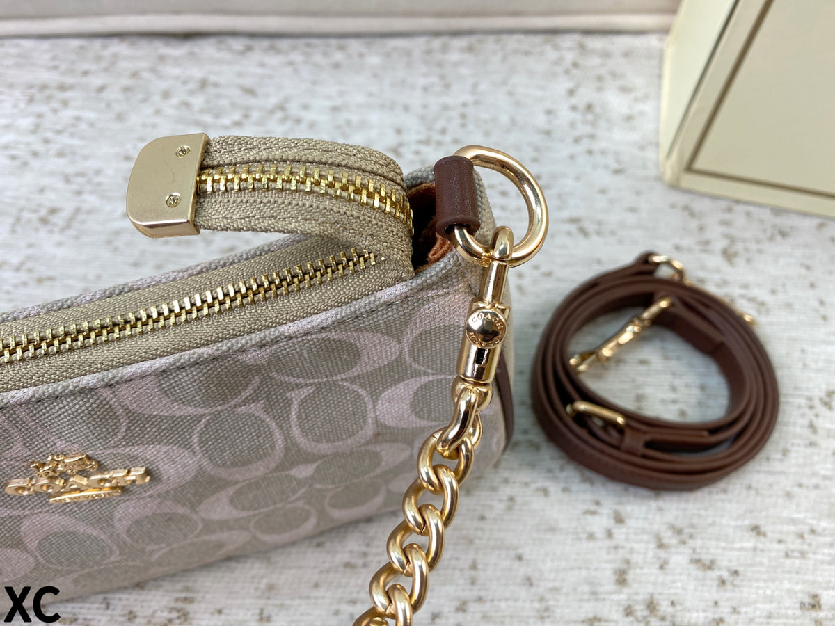 A close-up of a Coach zip pouch in the signature monogram canvas, featuring gold-tone hardware, a brown leather wristlet strap, and a gold logo on a textured light background.