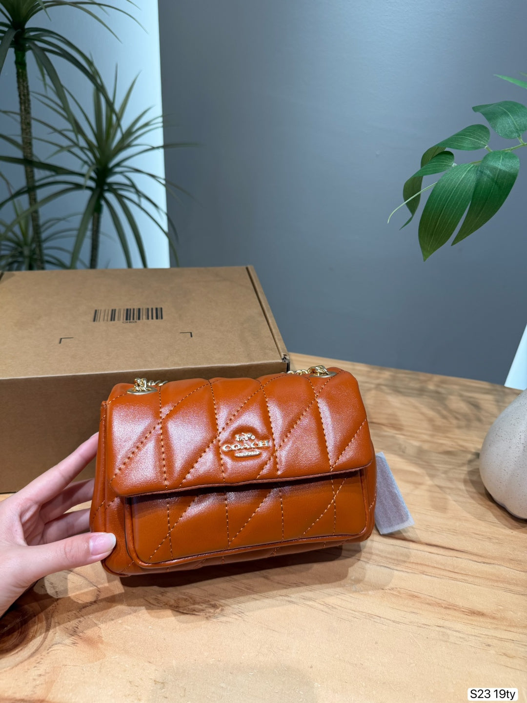 A hand holds a small quilted brown leather handbag in front of a cardboard box on a wooden table, with a gray wall and green plants in the background.