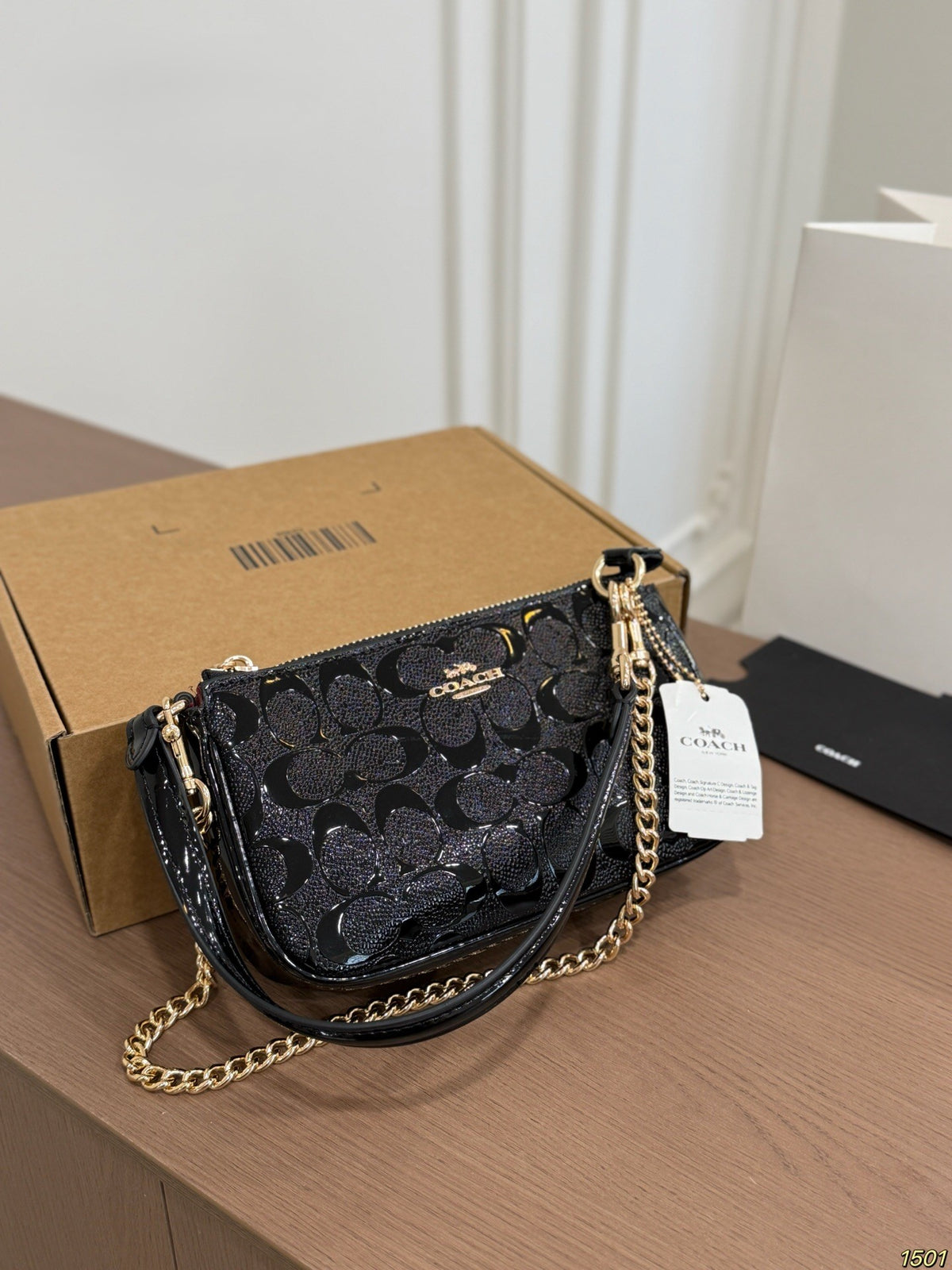 A Coach black glitter monogram shoulder bag with gold-tone chain strap, resting on a wooden table next to its brown cardboard box and a white product tag.