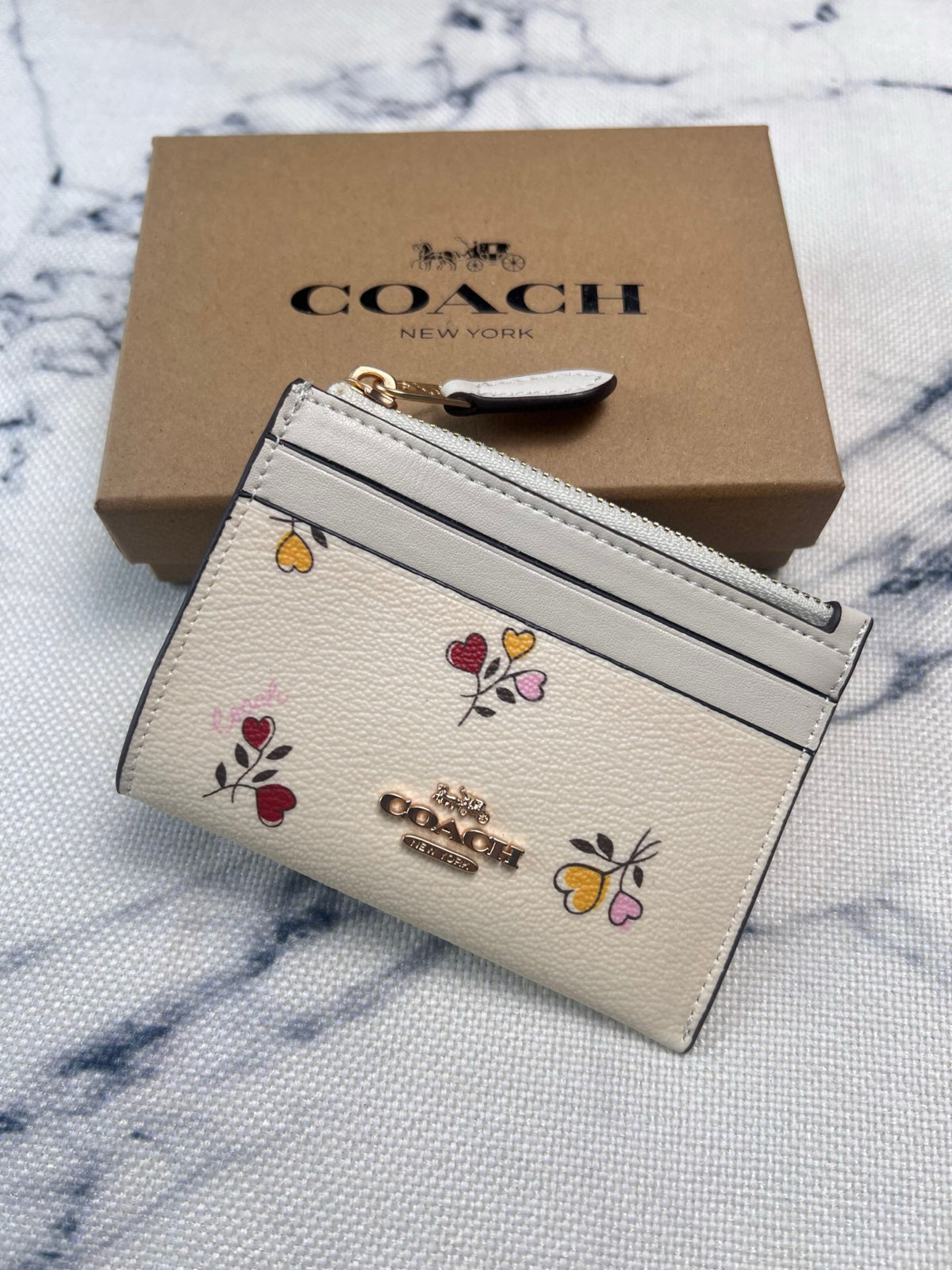 A Coach New York card holder wallet in textured cream leather with colorful floral and butterfly motifs, shown in front of its brown cardboard box on a white marble-patterned surface.