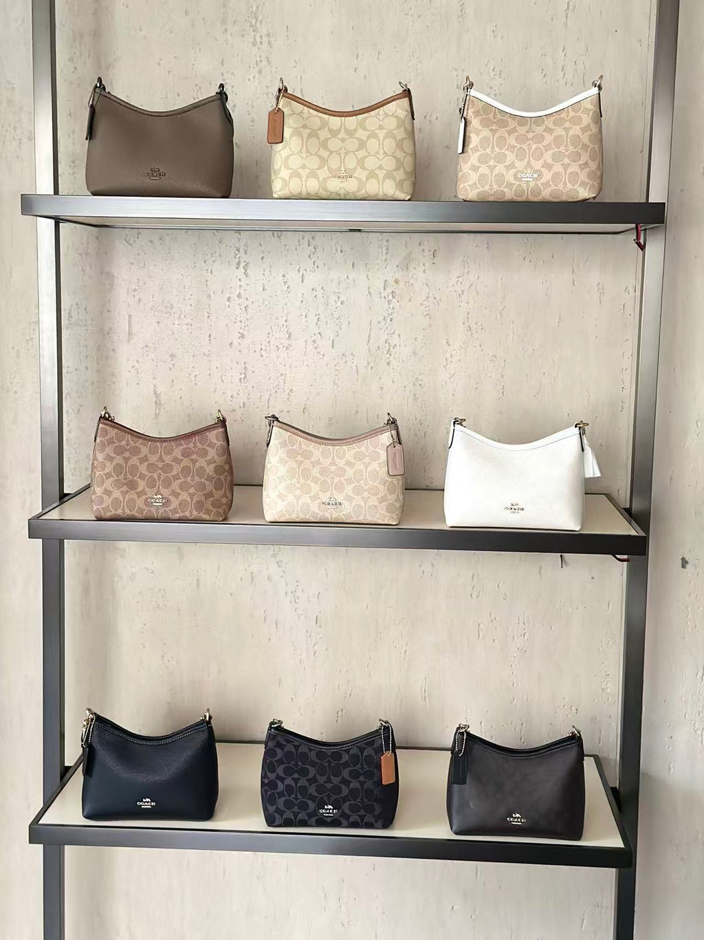 Nine Coach hobo shoulder bags in various colors and monogram patterns displayed on three shelves of a black metal rack against a textured light beige wall.
