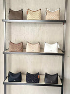 Nine Coach hobo shoulder bags in various colors and monogram patterns displayed on three shelves of a black metal rack against a textured light beige wall.