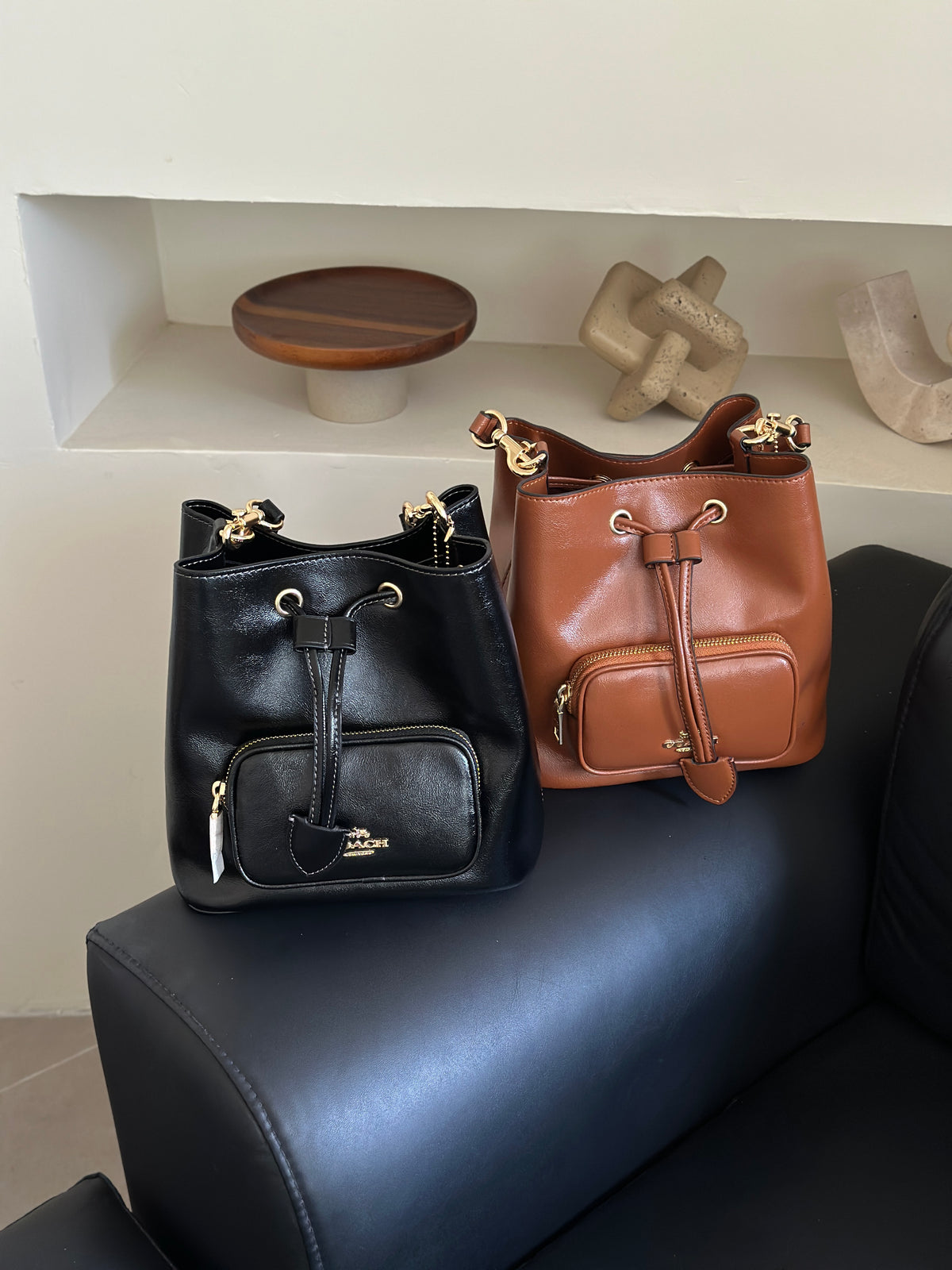 Two Coach leather bucket bags, one black and one tan-brown, displayed on a black leather sofa in a modern interior setting.