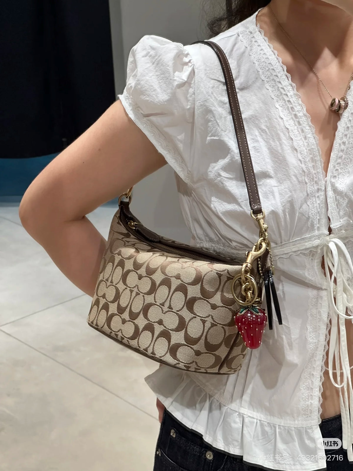 A person wearing a white blouse carries a Coach shoulder bag with the brand's signature monogram pattern in beige and brown. The bag features a brown leather strap, gold-tone hardware, and a red strawberry charm.