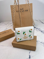 A Coach white leather shoulder bag with embroidered daisy flowers and green leaves, resting on a brown cardboard box with its signature Coach shopping bag and box in the background.