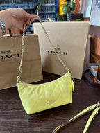 A Coach shoulder bag in light yellow patent leather with a gold chain strap, held by a hand in front of its brown Coach shopping bags on a dark wooden table.